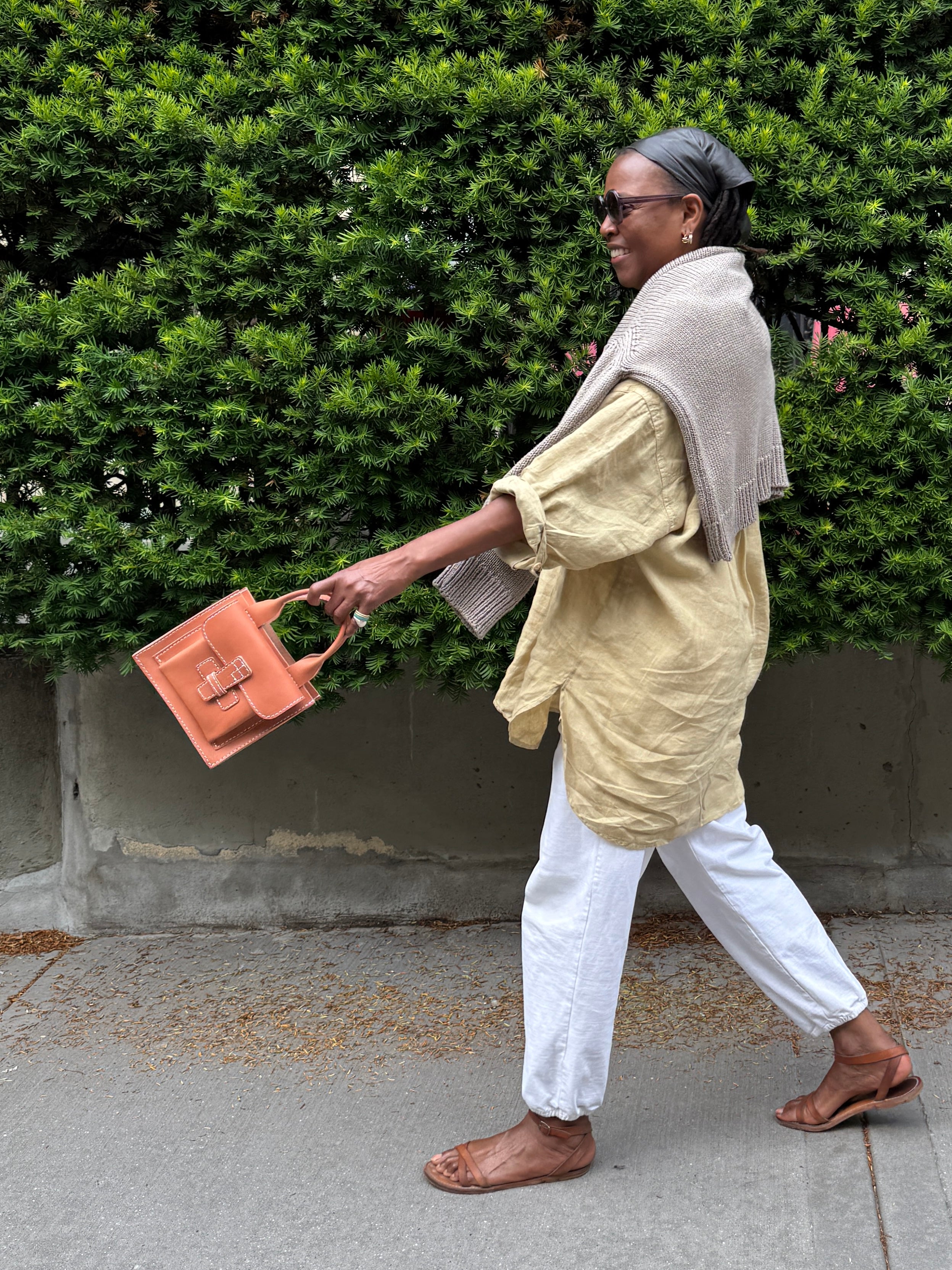 women walking and swinging a leather bag in front of foliage.