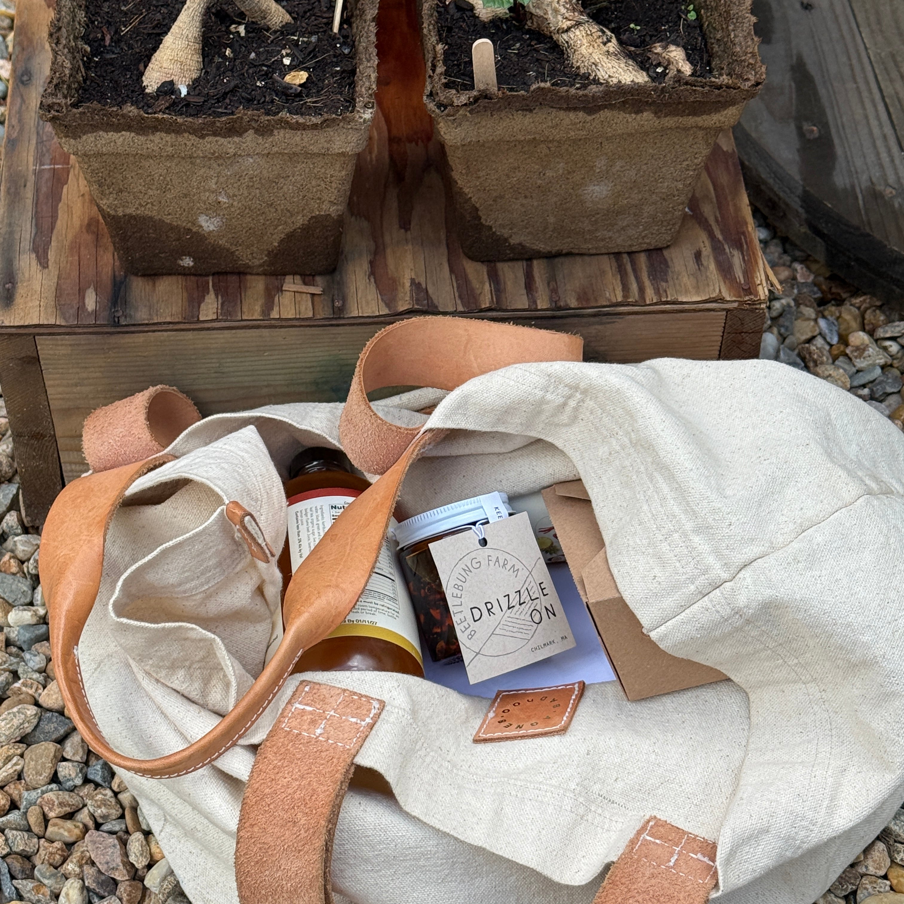 White tote bag with brown straps containing small bottles on a wooden surface with plants in the background.