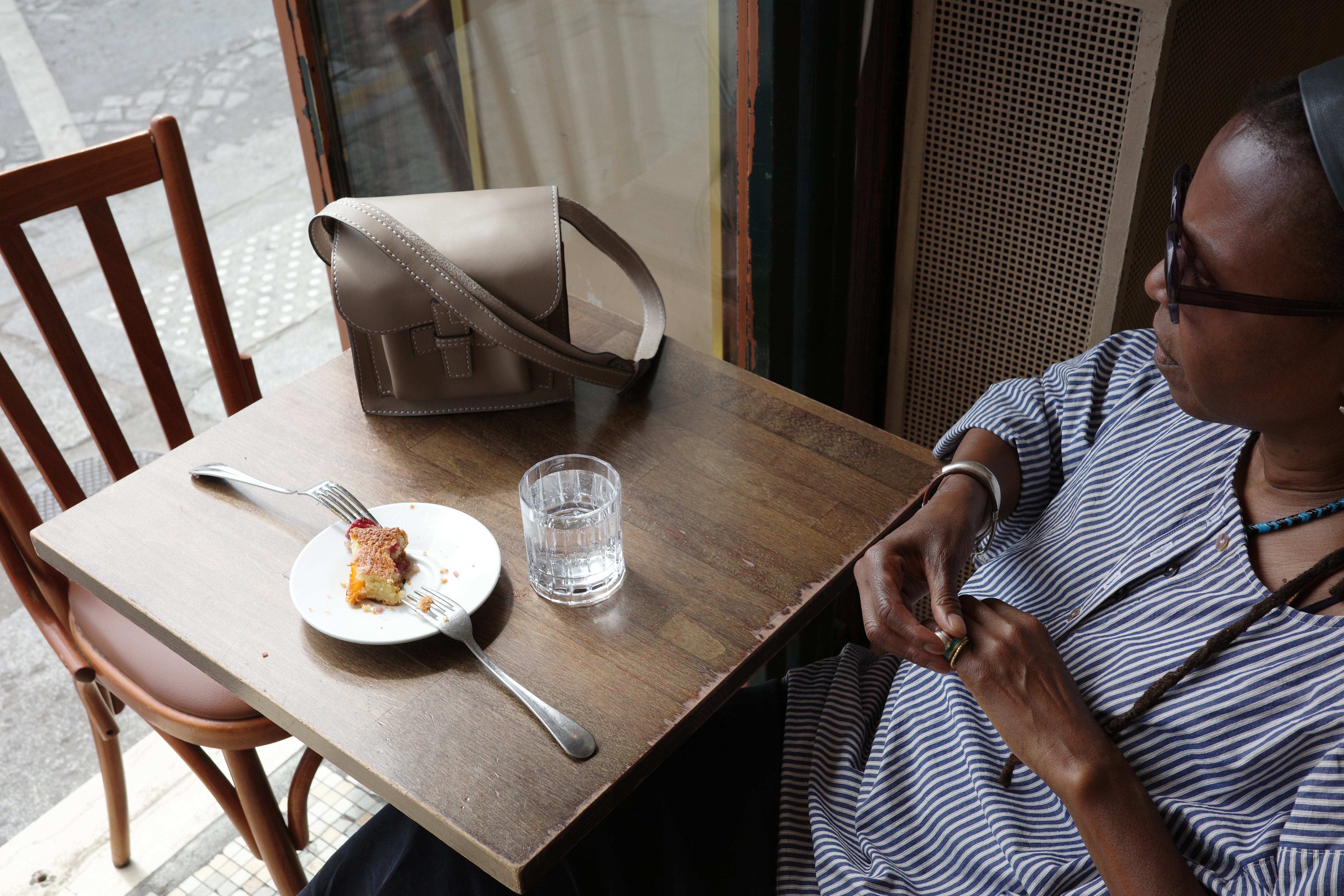 Agnes sitting at a table eating a slice of cake with a glass of water, next to a the sac .5 leather bag in grey. 