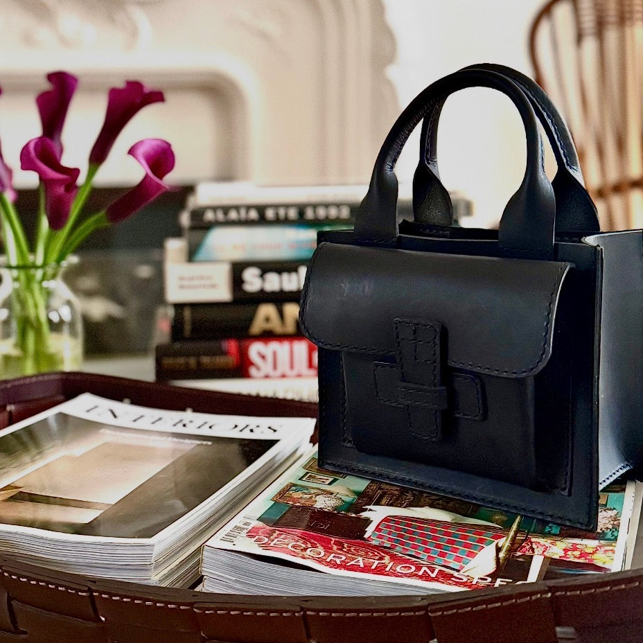 Black handbag on a table with magazines and a vase of flowers in the background