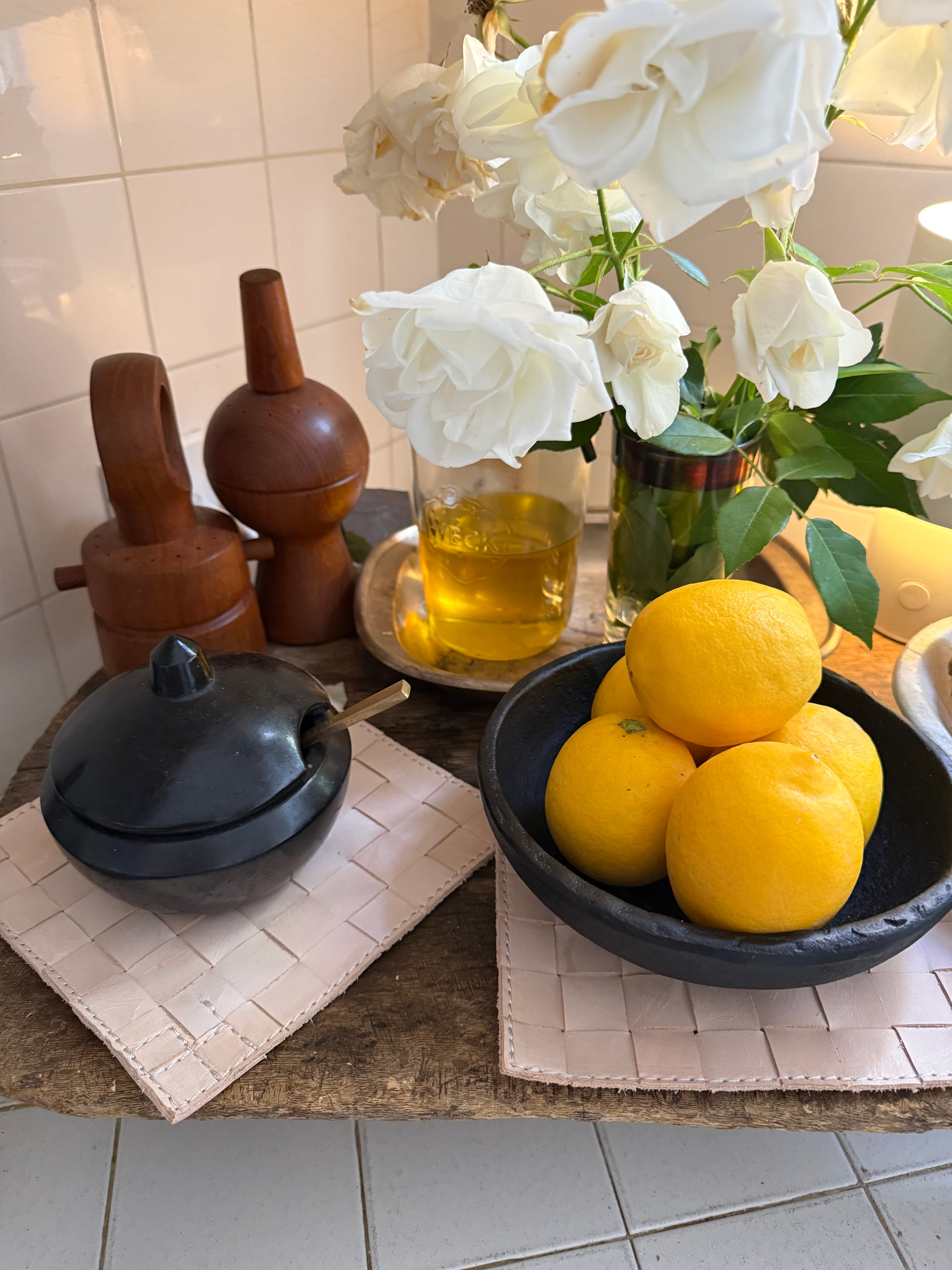 Bowl of lemons in a decorative bowl sitting on a leather trivet with flowers and candles in the background.