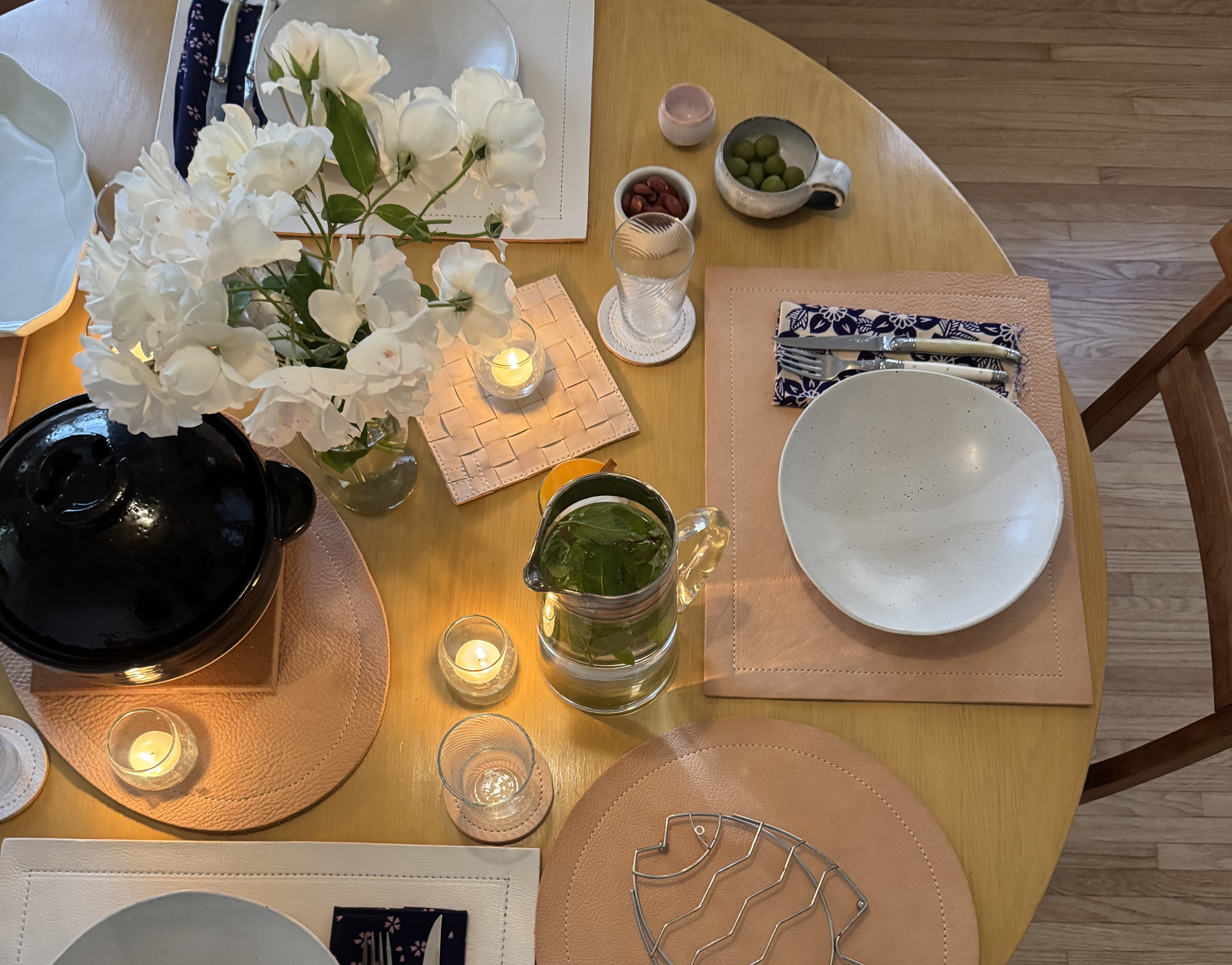 Overhead view of a round wooden dining table set for a cozy meal. The table features white bowls, folded patterned napkins with cutlery, leather placemats, and small lit candles. A vase of white flowers sits at the center alongside a glass pitcher filled with water and fresh mint leaves. Small dishes of olives and nuts are placed nearby, creating a warm, inviting atmosphere.