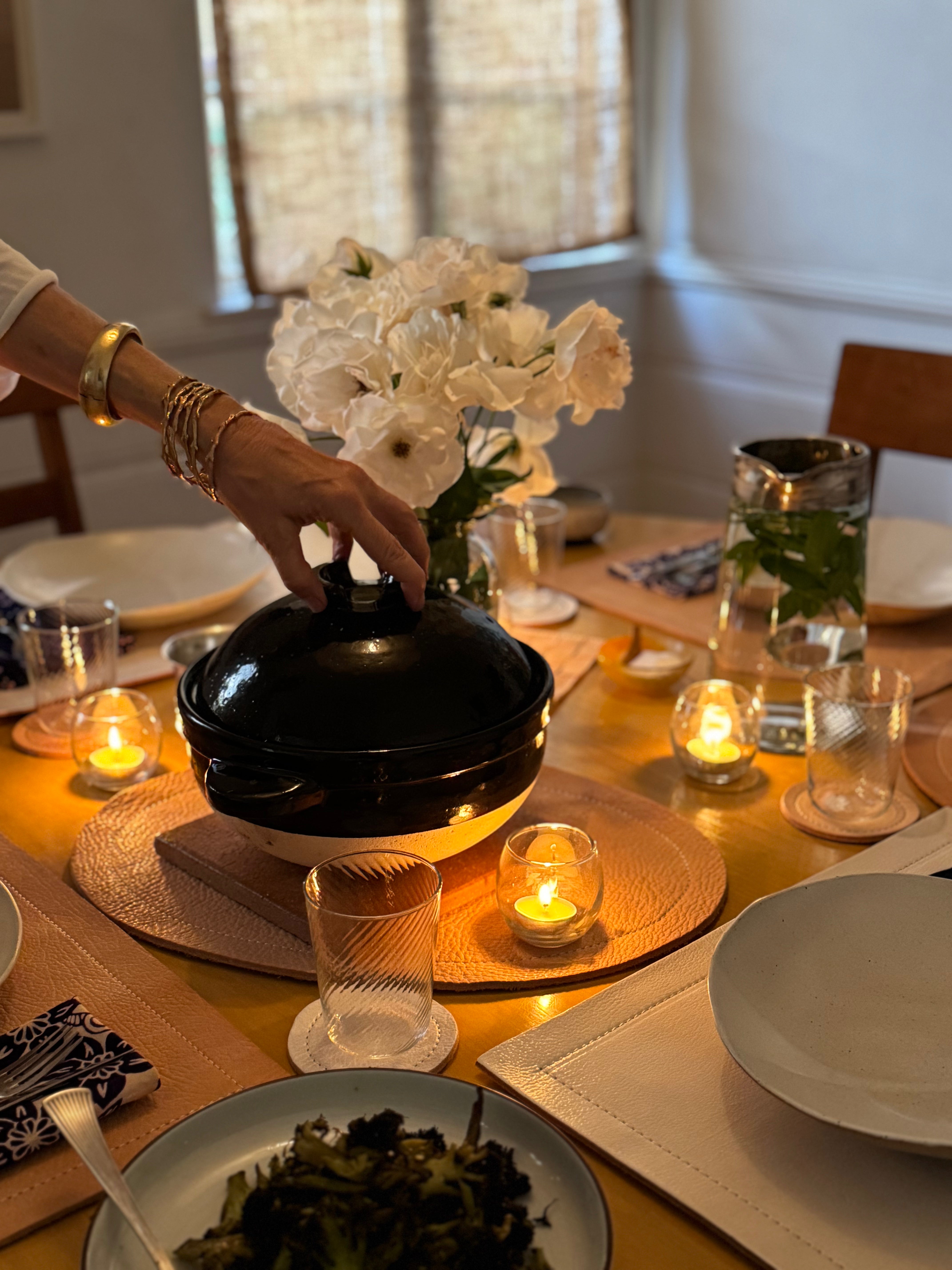 Dining table set with plates, glasses, and a vase of white flowers, with a person placing a black pot on the table.