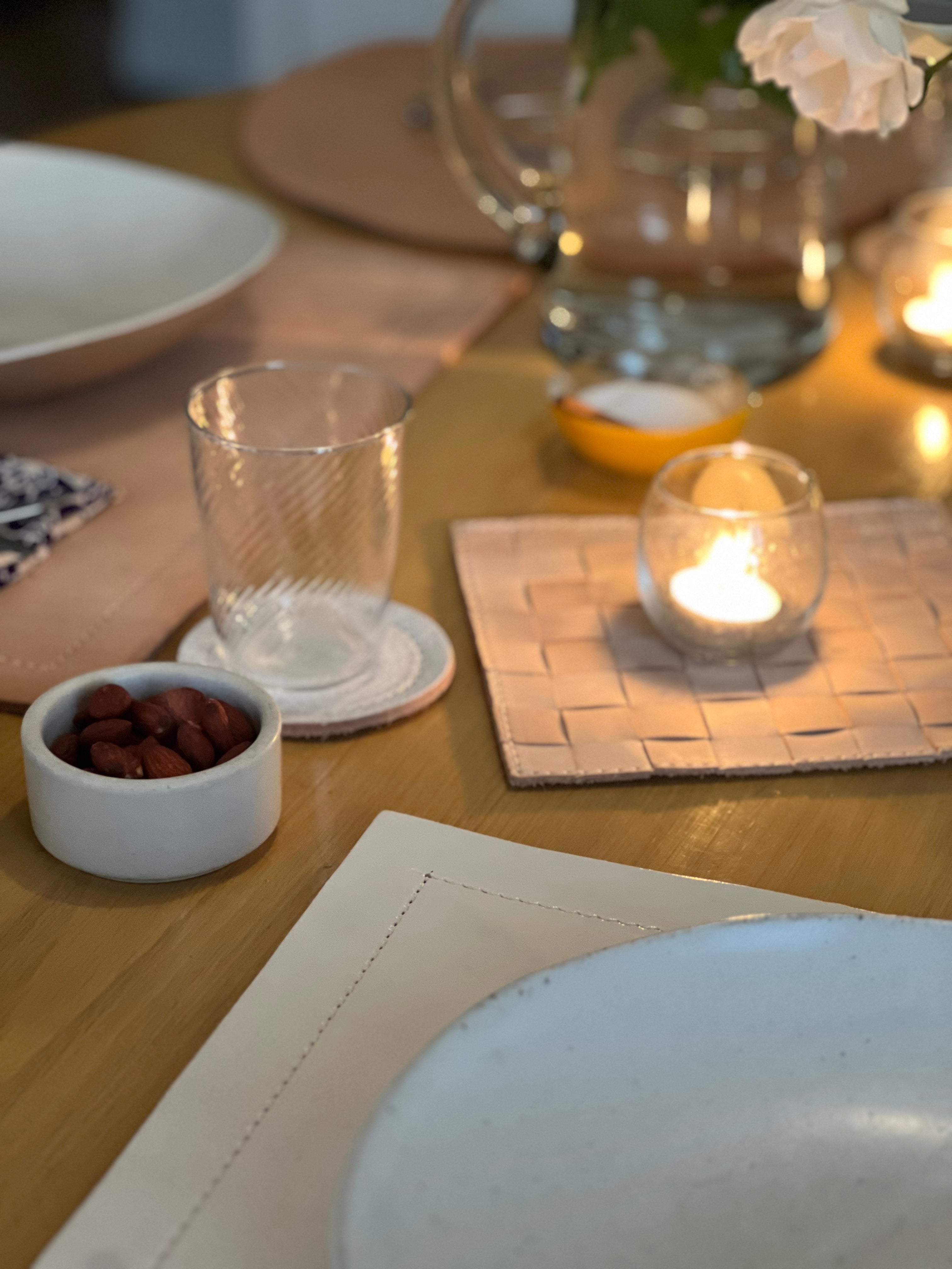 Close-up of a warmly lit dining table showing a textured glass on a round coaster, a small bowl of nuts, and a lit candle on a woven square mat. A white plate sits in the foreground, with a glass pitcher and flowers softly blurred in the background. 