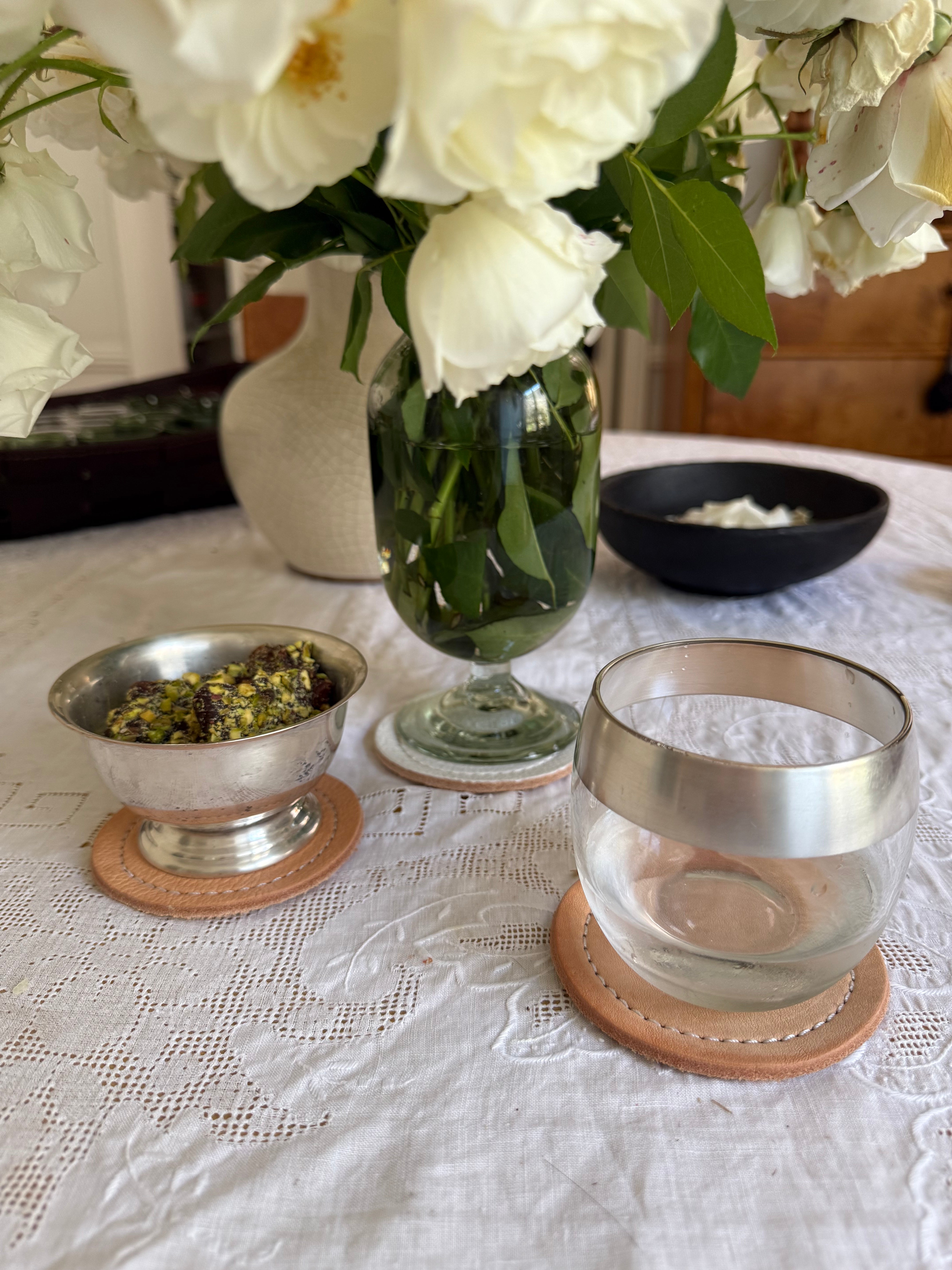 Tea-making setup with a glass teapot, silver bowls, and white flowers on a lace tablecloth.