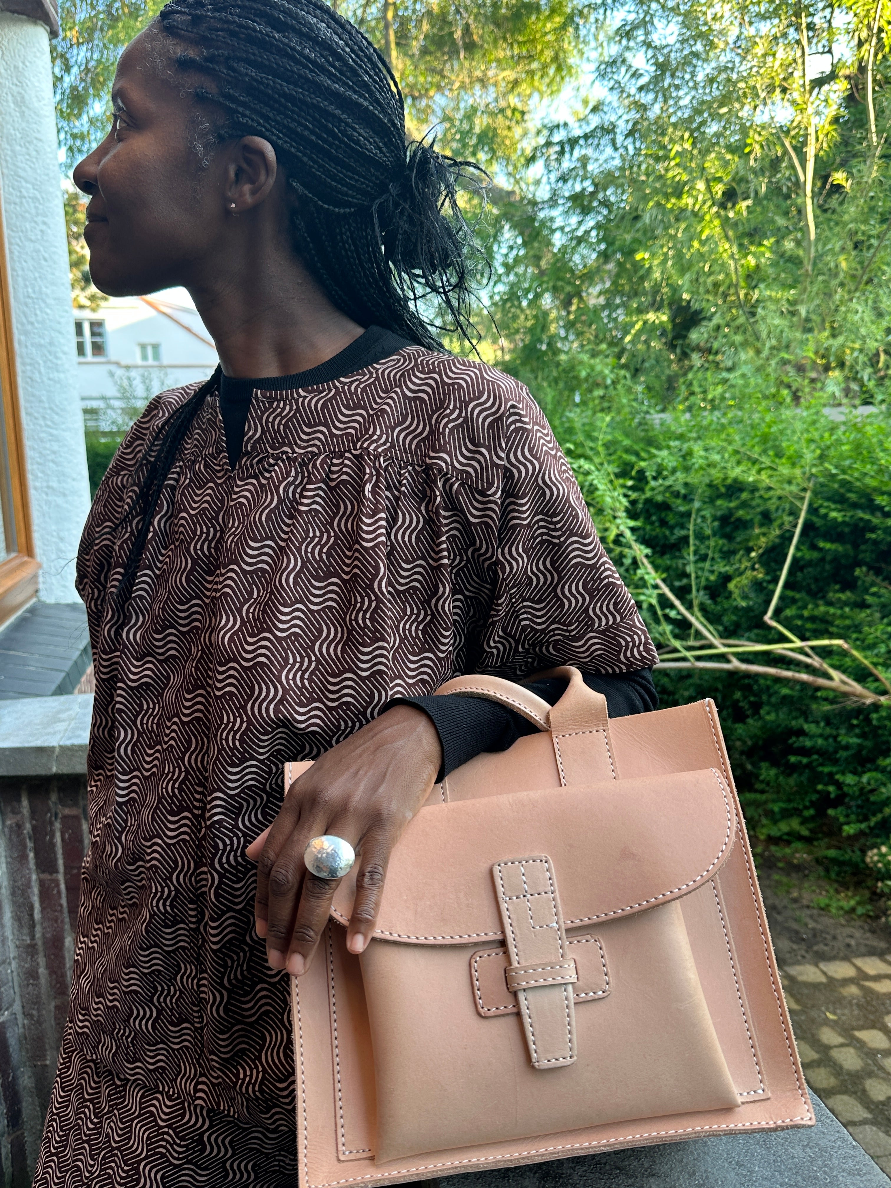 Person holding a leather handbag outdoors with greenery in the background