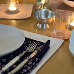 A warmly lit dining table set for a meal, featuring a white plate, fork, and knife resting on a dark floral napkin atop a leather placemat. Nearby are a textured glass on a coaster, a small metal bowl, and softly glowing candles that create a cozy, intimate atmosphere.
