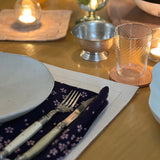 A warmly lit dining table set for a meal, featuring a white plate, fork, and knife resting on a dark floral napkin atop a leather placemat. Nearby are a textured glass on a coaster, a small metal bowl, and softly glowing candles that create a cozy, intimate atmosphere.