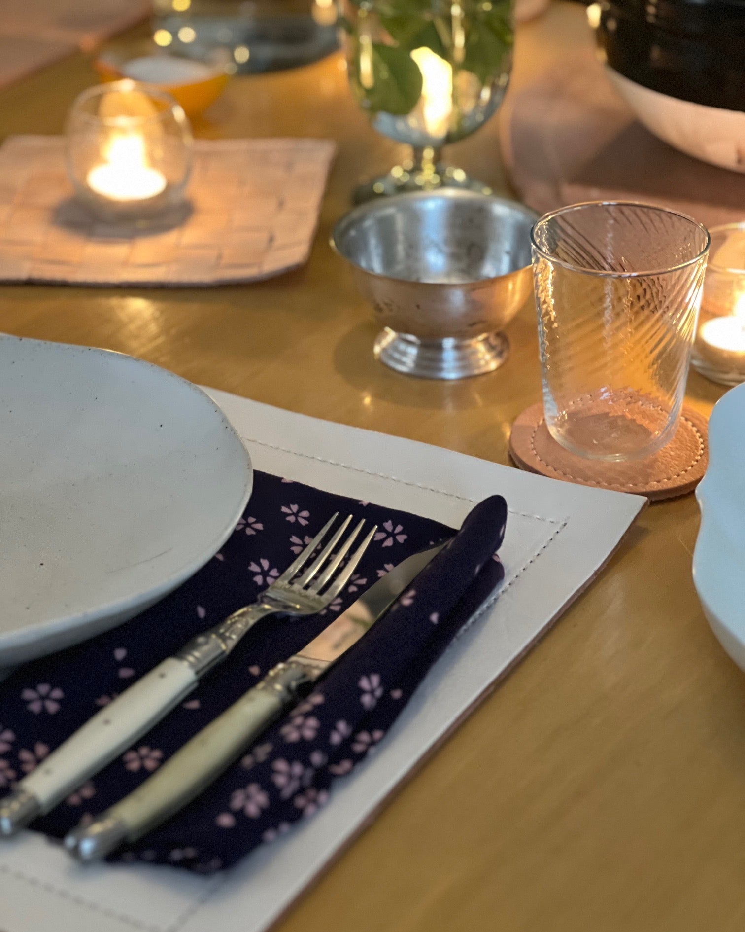 A warmly lit dining table set for a meal, featuring a white plate, fork, and knife resting on a dark floral napkin atop a leather placemat. Nearby are a textured glass on a coaster, a small metal bowl, and softly glowing candles that create a cozy, intimate atmosphere.